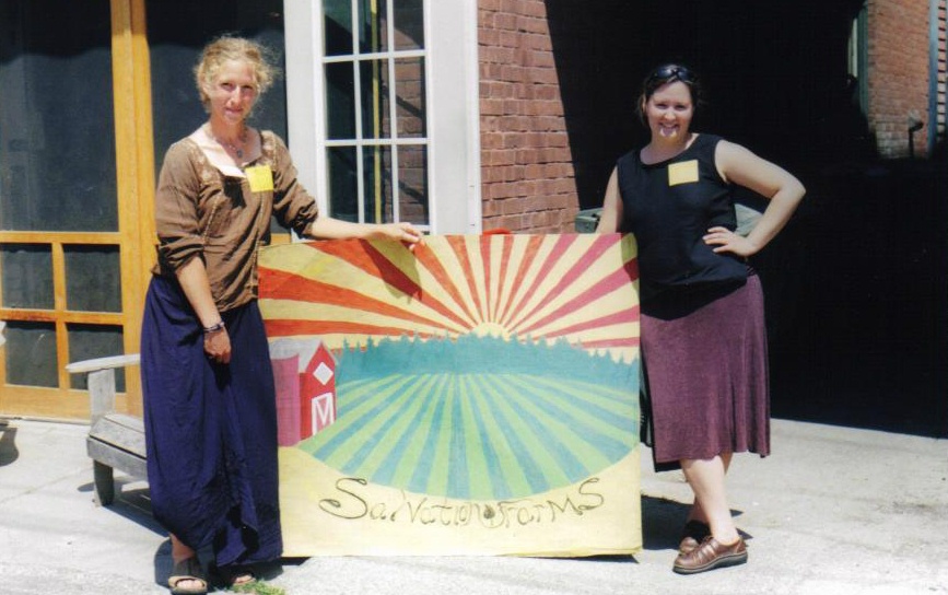 two women stand next to a painted sign that reads Salvation Farms. There is a red barn and green fields in the background of the sign